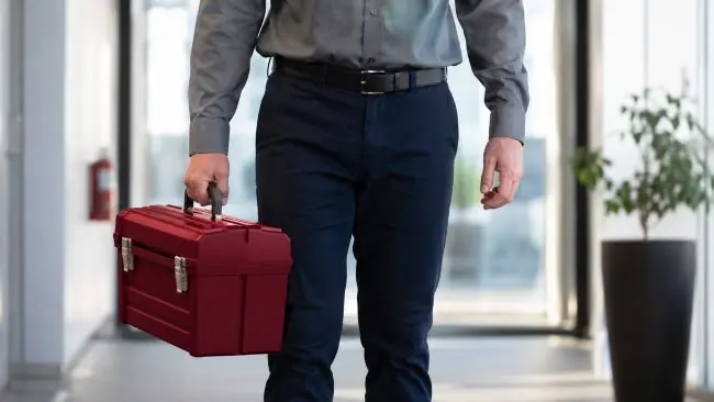 Ein Mann im Business Outfit steht im Gang eines Büros mit einem roten Werkzeugkoffer in der Hand als Symbol für Umsetzungsstärke